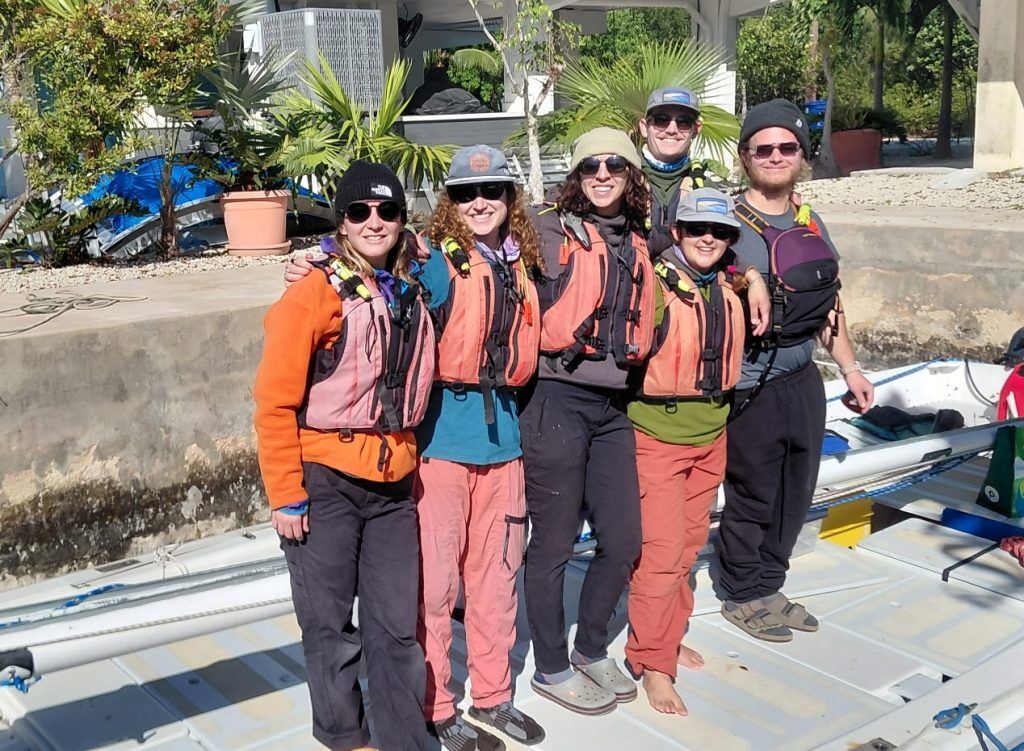 Here is a group of seven people posing for a photo. They are all wearing life vests and sunglasses, suggesting they are about to participate in a water activity. The background includes some plants and a building. The overall impression is that they are ready for an adventure.